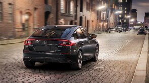 3/4 side rear view of black Hatchback taking a turn on a city street, in morning light.