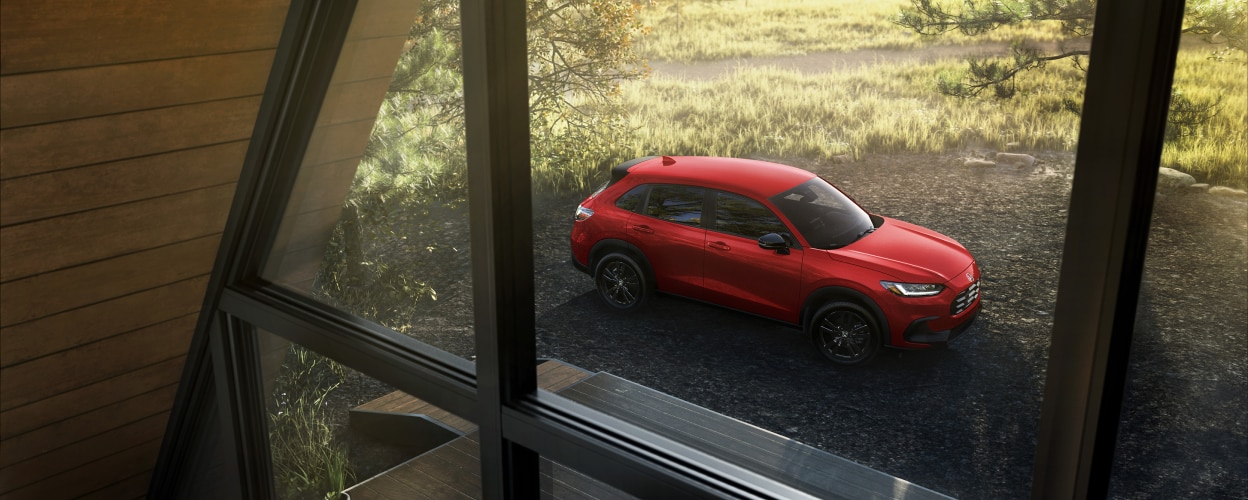 3/4 bird’s eye view of a red HR-V parked in the countryside, seen through the windows of an A-frame cottage. 