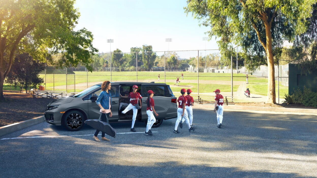 Mom and kids’ baseball team exiting a grey Odyssey parked near a baseball diamond.