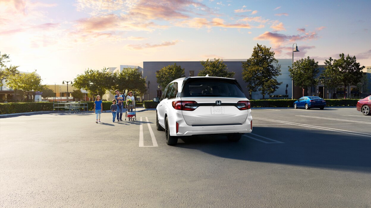 Rear view of grey Odyssey in empty parking lot. Family of walks toward it, pushing a grocery cart.