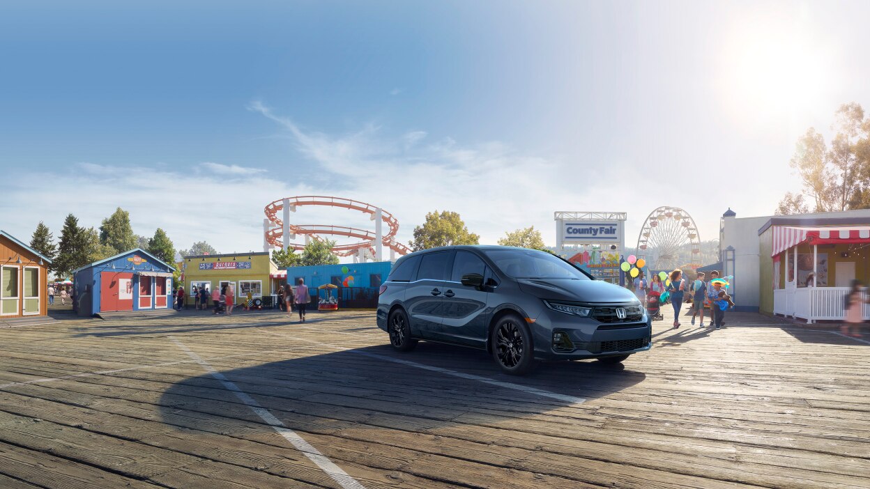 3/4 side front view of grey Odyssey parked in front of a county fair with roller coaster and ferris wheel int he background.