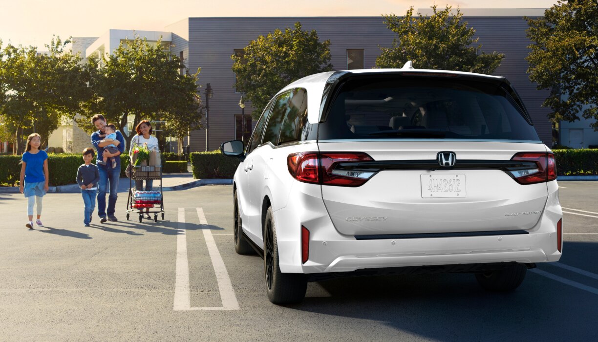 Rear view of grey Odyssey in empty parking lot. Family of walks toward it, pushing a grocery cart.