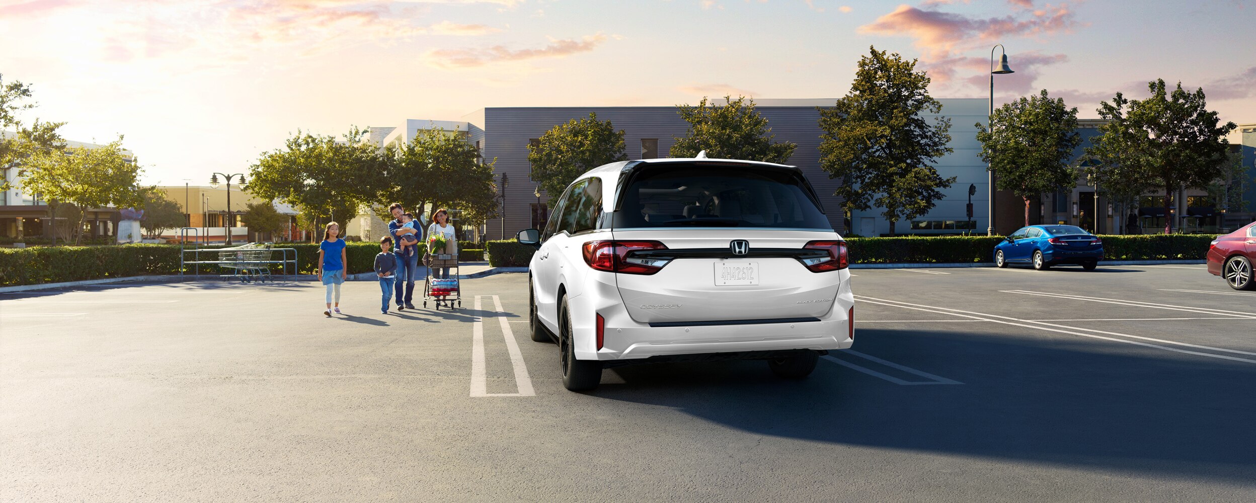 Rear view of grey Odyssey in empty parking lot. Family of walks toward it, pushing a grocery cart.