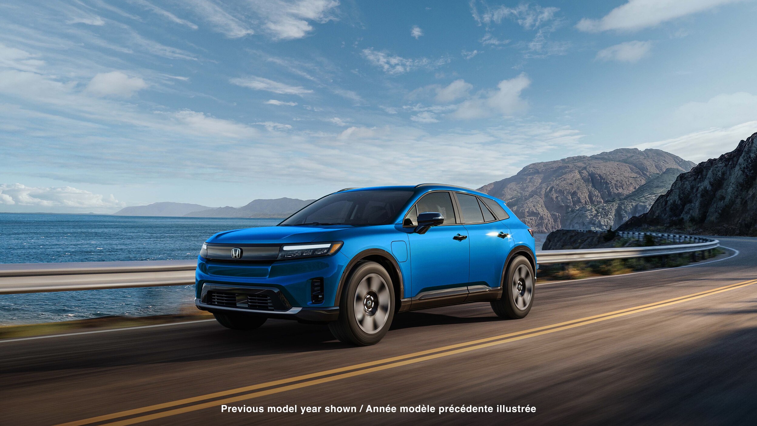Wide 3/4 front view of blue Prologue driving on coastal highway ith mountains in the background.