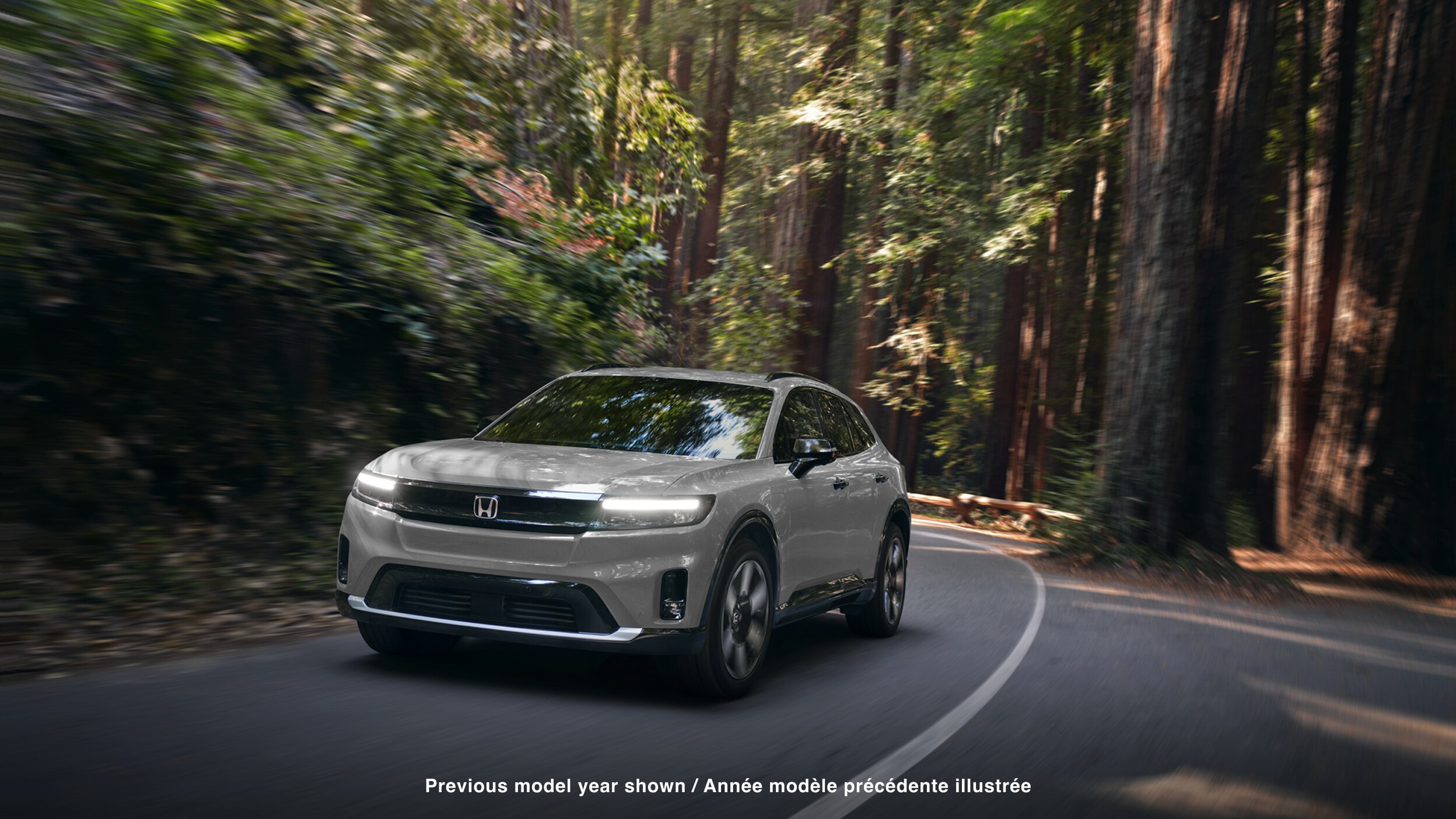 3/4 front view of white Prologue taking a turn on a paved road in a temperate forest.