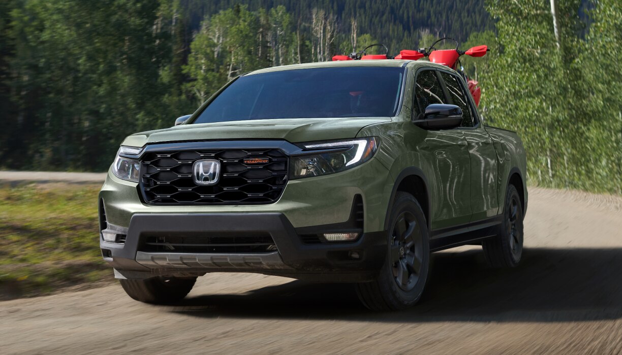3/4 front wide view of grey Ridgeline driving in on a tree-lined road. 