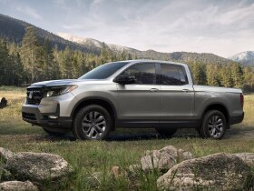 Side view of grey Ridgeline parked in an open grass field with temperate coniferous forest and mountains in the background. 